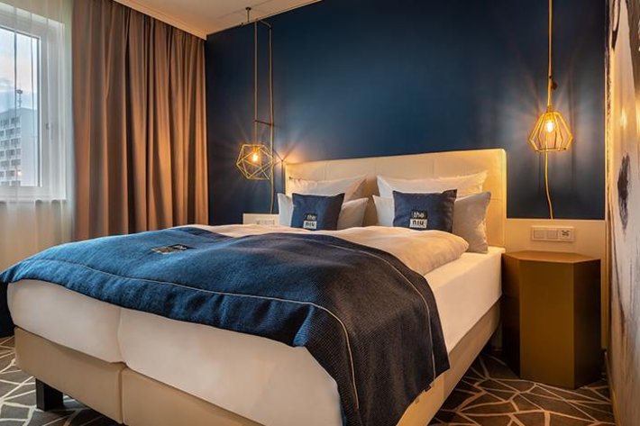 Modernes Hotelzimmer mit einem großen Doppelbett, blauen Wänden und stilvollen Pendelleuchten. Gemütliche Atmosphäre.