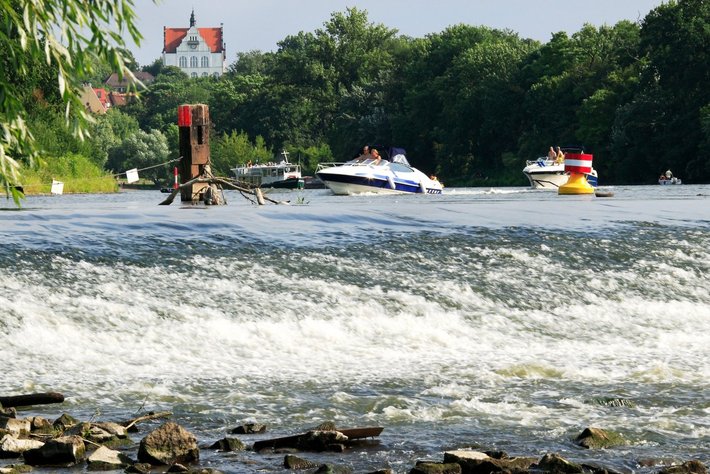 Hier wird ein alternativer Text für sehbehinderte Besucher angeboten:

„Bildunterschrift: Bootshaven am Fluss mit mehreren Motorsatkanäher an der Oberfläche. Im Hintergrund zeigt sich ein Gebäude mit steilem roten Dach. Turbulentes Wasser breitet sich im Vordergrund aus.“