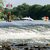 Hier wird ein alternativer Text für sehbehinderte Besucher angeboten:

„Bildunterschrift: Bootshaven am Fluss mit mehreren Motorsatkanäher an der Oberfläche. Im Hintergrund zeigt sich ein Gebäude mit steilem roten Dach. Turbulentes Wasser breitet sich im Vordergrund aus.“