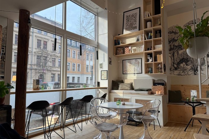Gemütliches Café mit großen Fenstern, die Blick auf eine Stadtstraße mit historischen Gebäuden bieten. Im Raum stehen zwei runde Tische mit Stühlen, eine gemütliche Sitzbank mit Kissen sowie Regale mit Büchern und Pflanzen.