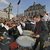 Große Orchesterprobe im Freien mit vielen Musikern, die Instrumente wie Streich-, Blas- und Schlaginstrumente spielen. Im Vordergrund ein Schlagzeuger, der auf einem großen Bass-Drum steht und mit Stöcken spielt.