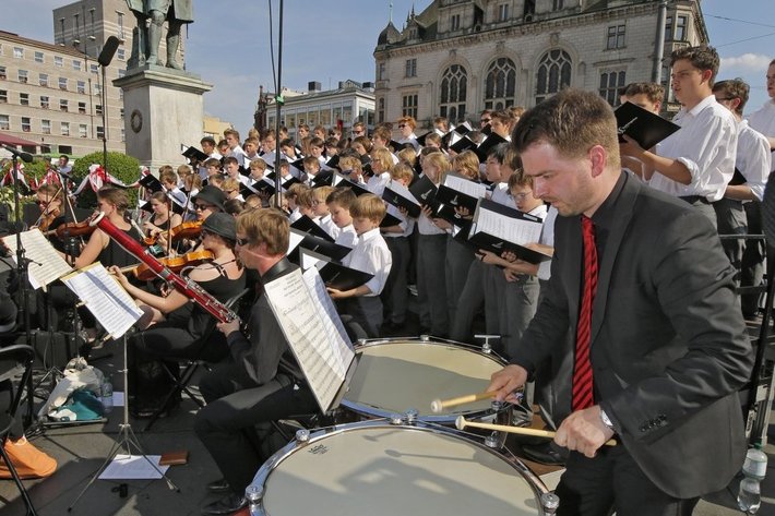 Große Orchesterprobe im Freien mit vielen Musikern, die Instrumente wie Streich-, Blas- und Schlaginstrumente spielen. Im Vordergrund ein Schlagzeuger, der auf einem großen Bass-Drum steht und mit Stöcken spielt.