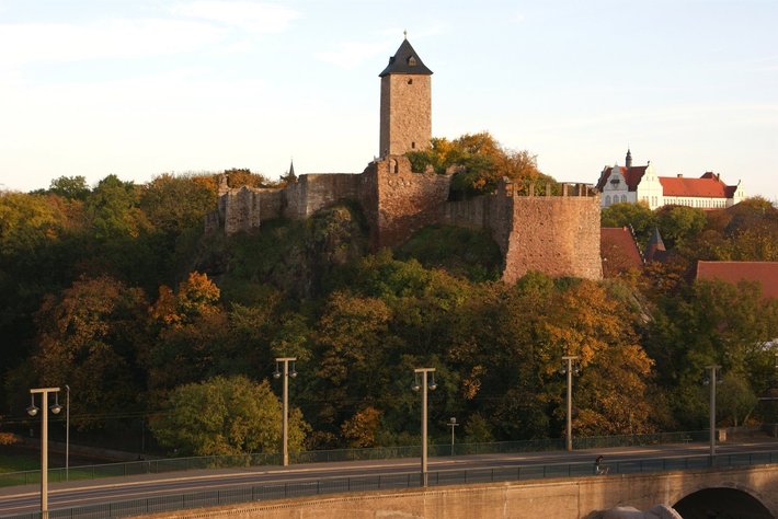 Diese Bild zeigt eine historische Burg auf einem Hügel mit grauen Steinmauern und einem schlanken Bergfried mit dunklerem Dach. Um die Burg liegen viele Bäume in herbstlichen Farben. ???????.