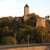 Diese Bild zeigt eine historische Burg auf einem Hügel mit grauen Steinmauern und einem schlanken Bergfried mit dunklerem Dach. Um die Burg liegen viele Bäume in herbstlichen Farben. ???????.