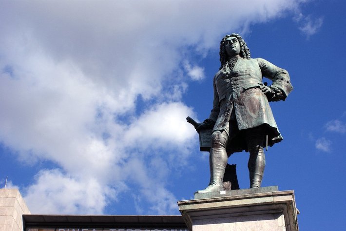 Bronzestatue eines historischen Mannes in barocker Kleidung, der mit einer Hand auf einem Stock oder Zepter steht, auf einem Sockel vor blauem Himmel mit Wolken.