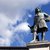 Bronzestatue eines historischen Mannes in barocker Kleidung, der mit einer Hand auf einem Stock oder Zepter steht, auf einem Sockel vor blauem Himmel mit Wolken.