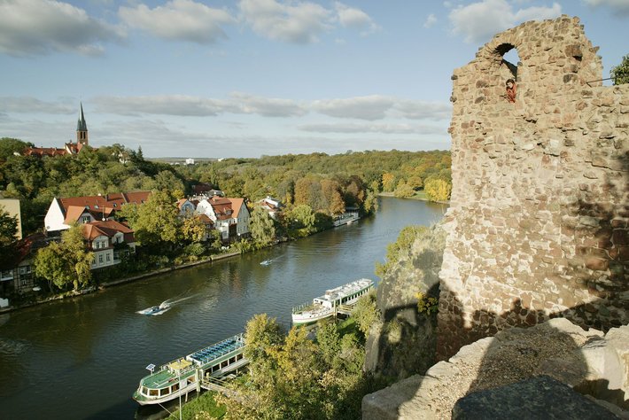 Blick von einer alten Burgruine auf eine Flusslandschaft mit Booten, darunter ein grünes Ausflugsschiff und ein kleines Motorboot. Im Hintergrund eine malerische Stadt mit Fachwerkhäusern und Bäumen im Herbstlicht.