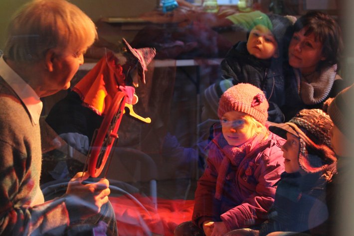 In dieser Abbildung spielt eine ältere Person Geige vor einem Publikum aus Kindern. Die Kinder schauen attent und gespannt zu der performance, teilweise eingemummt. Gemütliche Atmosphäre mit festlicher, warmgetönter Beleuchtung.