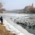 In dieser winterlichen Szene stehen am Ufer eines partiell zugefrorenen Flusses einige Personen und beobachten eine große Gruppe von Enten. Der Fluss liegt sicher in der Nähe eines Brücken- und Gebäudehintergrunds, aber im Fokus sind die Vögel und die zugeschneite Landschaft.