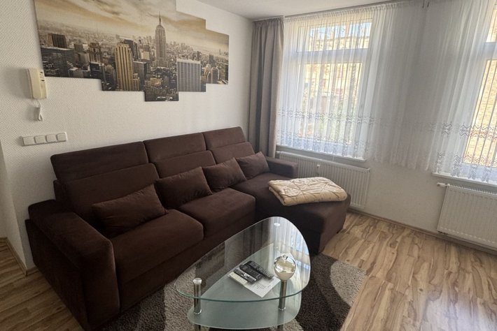 Gemütliches Wohnzimmer mit braunem Sofa und Glas-Couchtisch in der Mitte, daneben eine Uhr. An der Wand ein großes Bild einer Stadt mit Wolkenkratzern, darunter ein Heizkörper. Großes Fenster mit Vorhang.