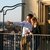 Ein Mann und eine Frau stehen auf einem Balkon mit Aussicht auf eine Stadt. Der Mann zeigt mit dem Arm in die Ferne, während beide Gläser in den Händen halten und die Skyline betrachten.