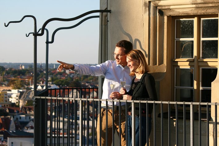 Ein Mann und eine Frau stehen auf einem Balkon mit Aussicht auf eine Stadt. Der Mann zeigt mit dem Arm in die Ferne, während beide Gläser in den Händen halten und die Skyline betrachten.