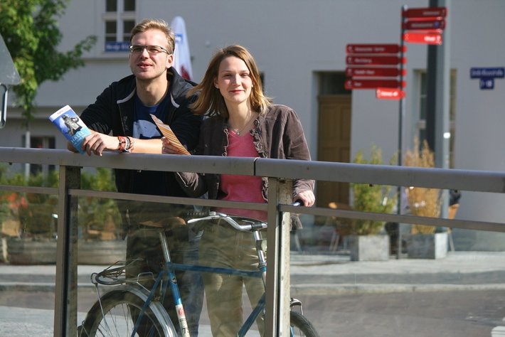 Ein junger Mann und eine junge Frau lehnen lächelnd an einem Geländer. Daneben steht ein Fahrrad. Beide wirken entspannt. Die Frau trägt einen dicken Mantel, der Mann einen Jackenpulli mit Kapuze.