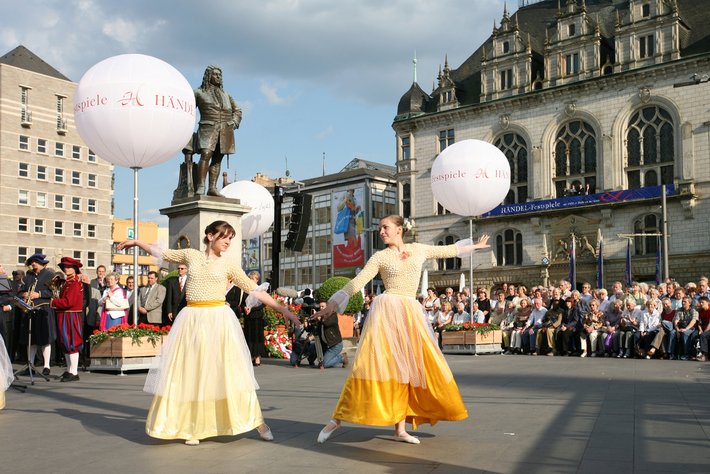 Zwei Frauen in traditioneller Tracht tanzen vor Publikum auf einem öffentlichen Platz, jede hält einen großen weißen Luftballon mit der Aufschrift „München“.