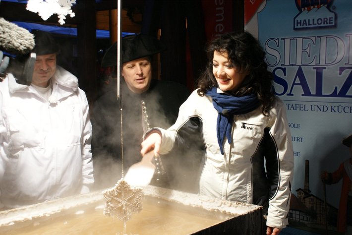 Eine Frau in warmer Kleidung gießt geschmolzenes Salz in eine flache Form, während zwei Männer in historischen Gewändern zuschauen. Im Hintergrund steht ein Schild mit der Aufschrift „Siedendes Salz“.