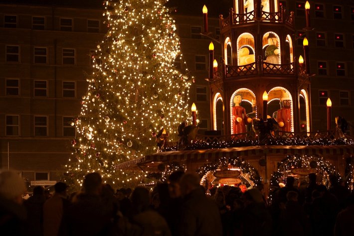 Ein wunderschön geschmückter Weihnachtsbaum, der mit kleinen Lichtern erleuchtet ist, steht im Mittelpunkt. Im Hintergrund ist eine festlich beleuchtete markante Gebäude-Silhouette mit Balkonen und weiteren Lichtern sowie verzierten Stützpfeilern zu sehen. Die Szene wirkt festlich und weihnachtlich und wird von Menschen durchlaufen.