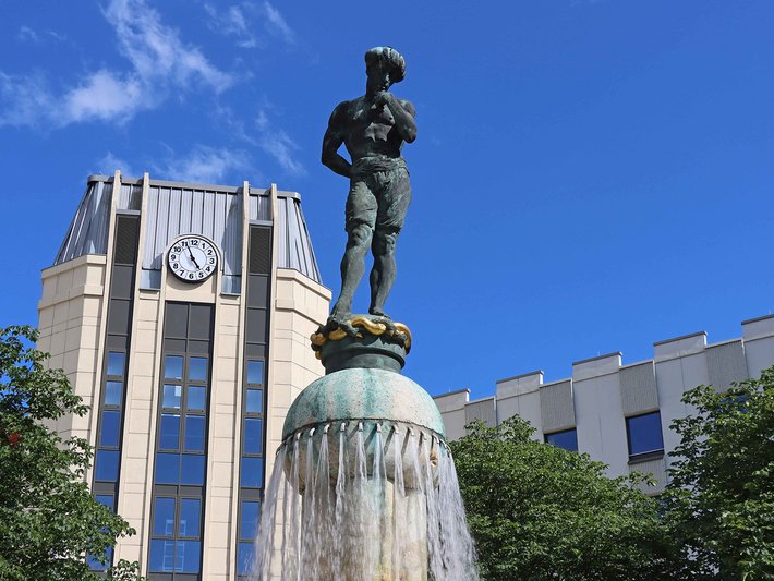 Bronzestatue eines Soldaten, der auf einem Brunnen mit Wasserfontäne steht, vor einem historischen Gebäude mit Uhr und klassischer Architektur.