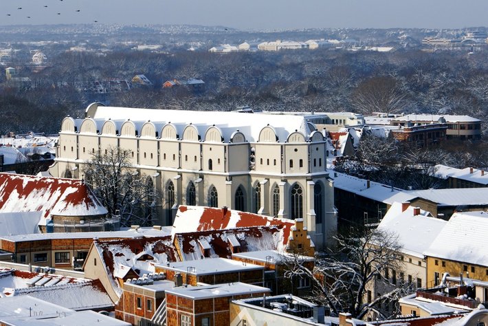 Diese Winterlandschaft zeigt eine überwölbte, große weiße oder hell beeinte Kirche mit vielen architektonischen Details wie Rundfenstern und symmetrischem Aufbau. Die Kirche ist von mit Schnee bedeckten Häusern mit rotbraunen Dächern umgeben. Die Szene wirkt still und winterlich.