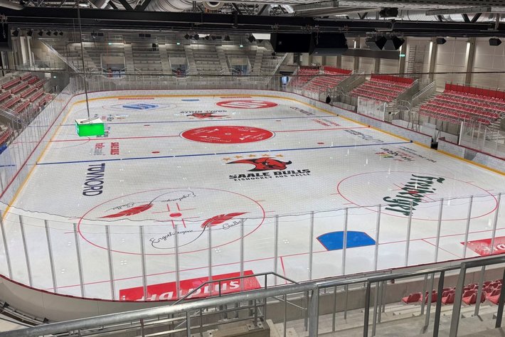 Leere Eishalle mit markiertem Spielfeld, darunter das Logo „Sharks“ und die Aufschrift „Halle Bulle“. Rote, blaue und grüne Werbeflächen auf dem Eis. Leere Tribünen mit roten Sitzen im Hintergrund.