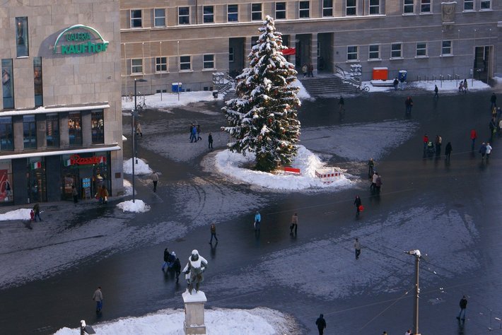 In diesem Großstadtplatz steht ein großer, mit Schnee bedeckter Weihnachtsbaum. Menschen gehen um ihn herum, die Fußwege sind teilweise vereist und eisglatt. Festliche Atmosphäre mit Weihnachtsstimmung in einer Innenstadtumgebung.
