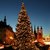 Ein festlich geschmückter, großer Weihnachtsbaum ist mit Lichterketten erleuchtet, umgeben von historischen Gebäuden mit Türmchen. Der Himmel ist dunkelblau, und es herrscht eine gemütliche u sowie weihnachtliche Stimmung vor.