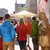 In dieser Abbildung stehen mehrere Personen in einer Gruppe in einer Stadtstraße. Ein Guide erklärt vermutlich Sehenswürdigkeiten. Rechts befindet sich eine goldfarbene Statue mit zwei historienreichen Figuren. Der Hintergrund zeigt eine altmodische Fachwerkfassade und besonders prächtige Architektur. (50 Wörter)