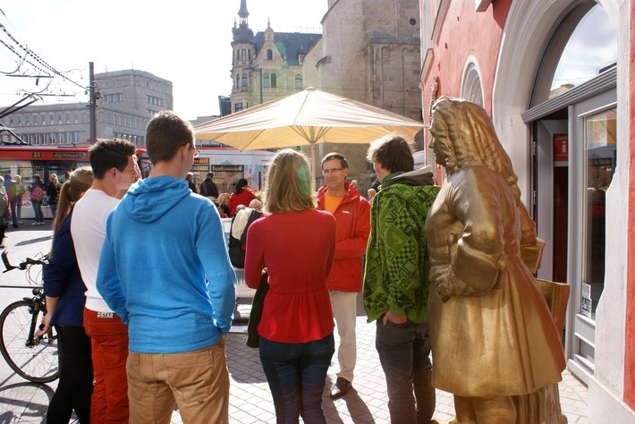 In dieser Abbildung stehen mehrere Personen in einer Gruppe in einer Stadtstraße. Ein Guide erklärt vermutlich Sehenswürdigkeiten. Rechts befindet sich eine goldfarbene Statue mit zwei historienreichen Figuren. Der Hintergrund zeigt eine altmodische Fachwerkfassade und besonders prächtige Architektur. (50 Wörter)