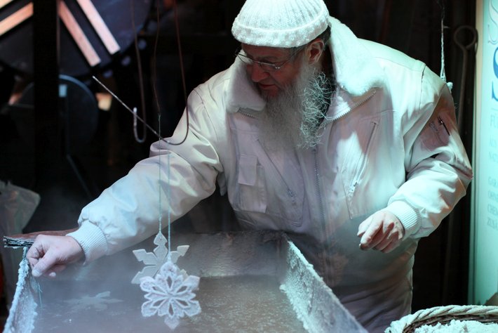 Een älterer Mann trägt eine weiße Robe und einen Hut. Er hält ein glitzerndes, sternförmiges Schneeflockendesign, das er vermutlich auf einen Sektkuchen platziert. Die Szene wirkt festlich und ritualhaft.